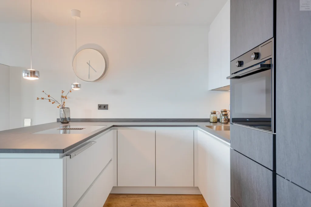 Moderne L-vormige keuken met witte en grijze kasten, houten vloer en ronde klok aan de muur.