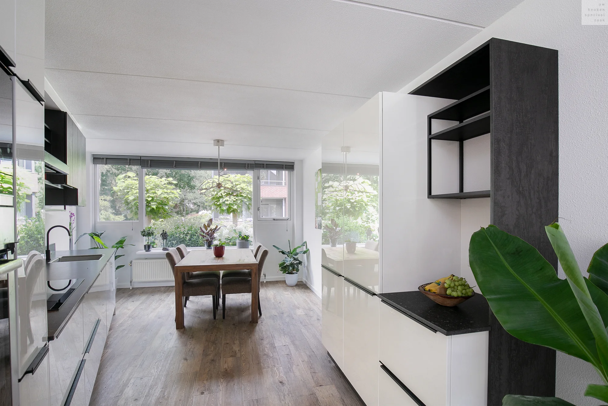 Moderne keuken met zwarte en witte kasten, houten eettafel, grote ramen en planten.