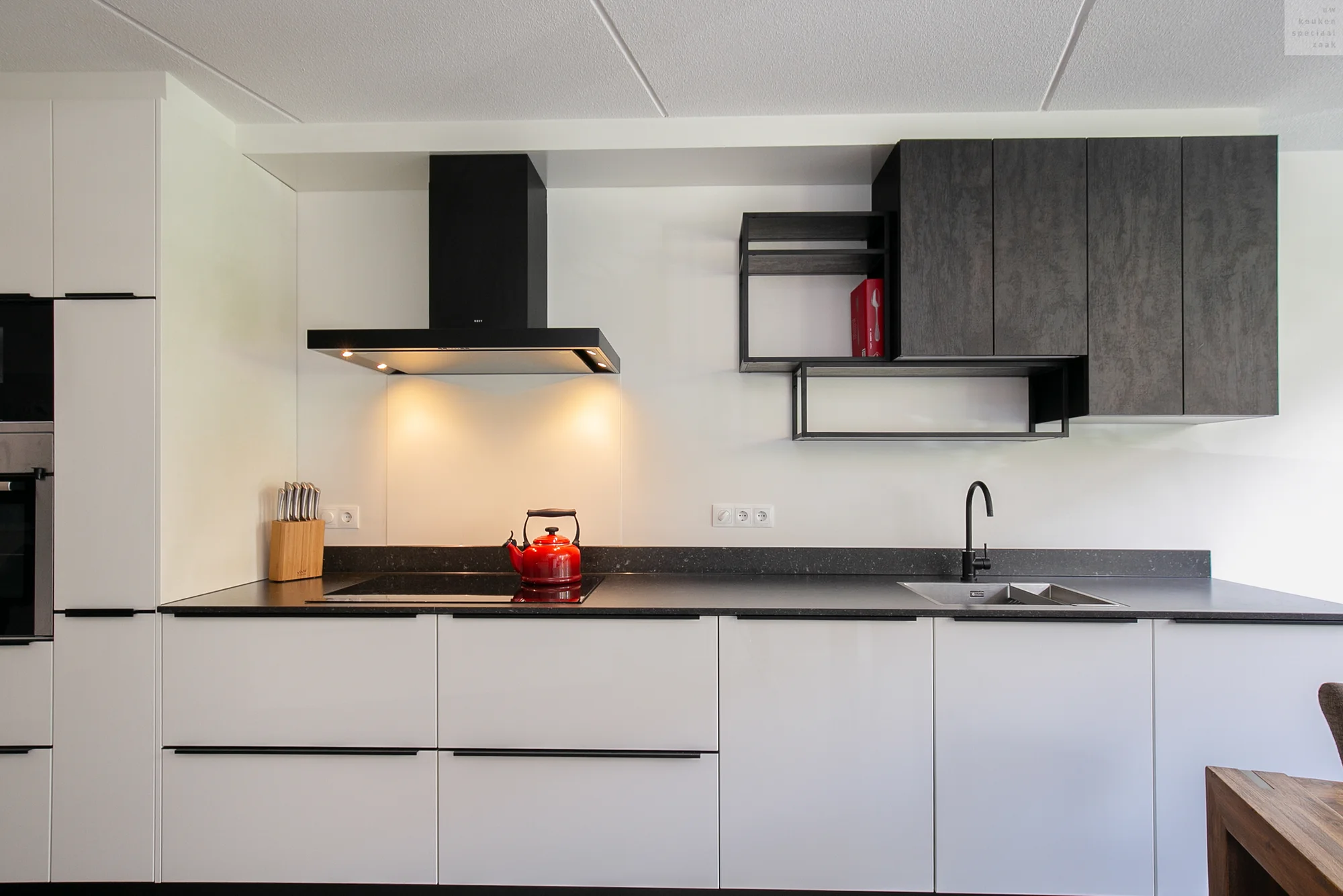 Moderne keuken met matte witte onderkasten, zwarte handgrepen, donkere bovenkasten en een zwarte afzuigkap