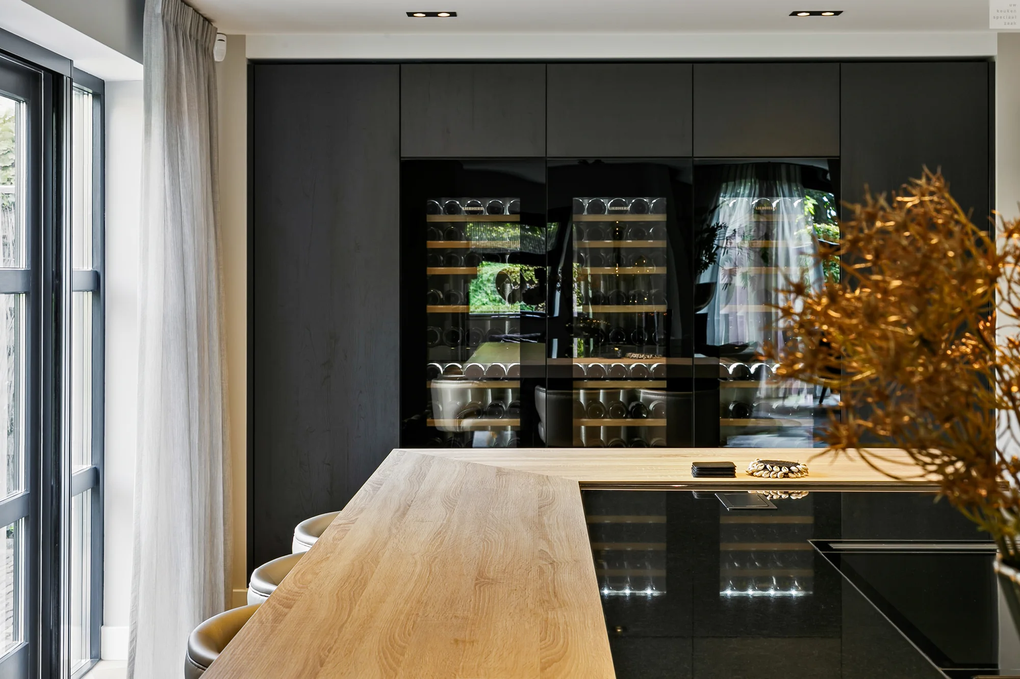 Moderne keuken met houten tafel, zwarte kast met wijnflessen en grote ramen met lichte gordijnen