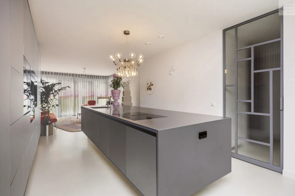 Moderne keuken met lang grijs keukeneiland, kookplaat, kroonluchter, grote ramen en zithoek met plant