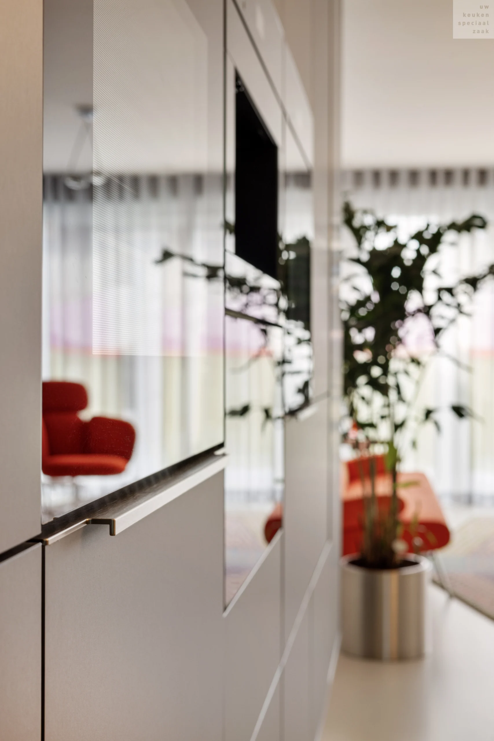 Moderne keuken met glanzende kast, metalen handgreep, grote ramen, groene plant en rood meubelstuk