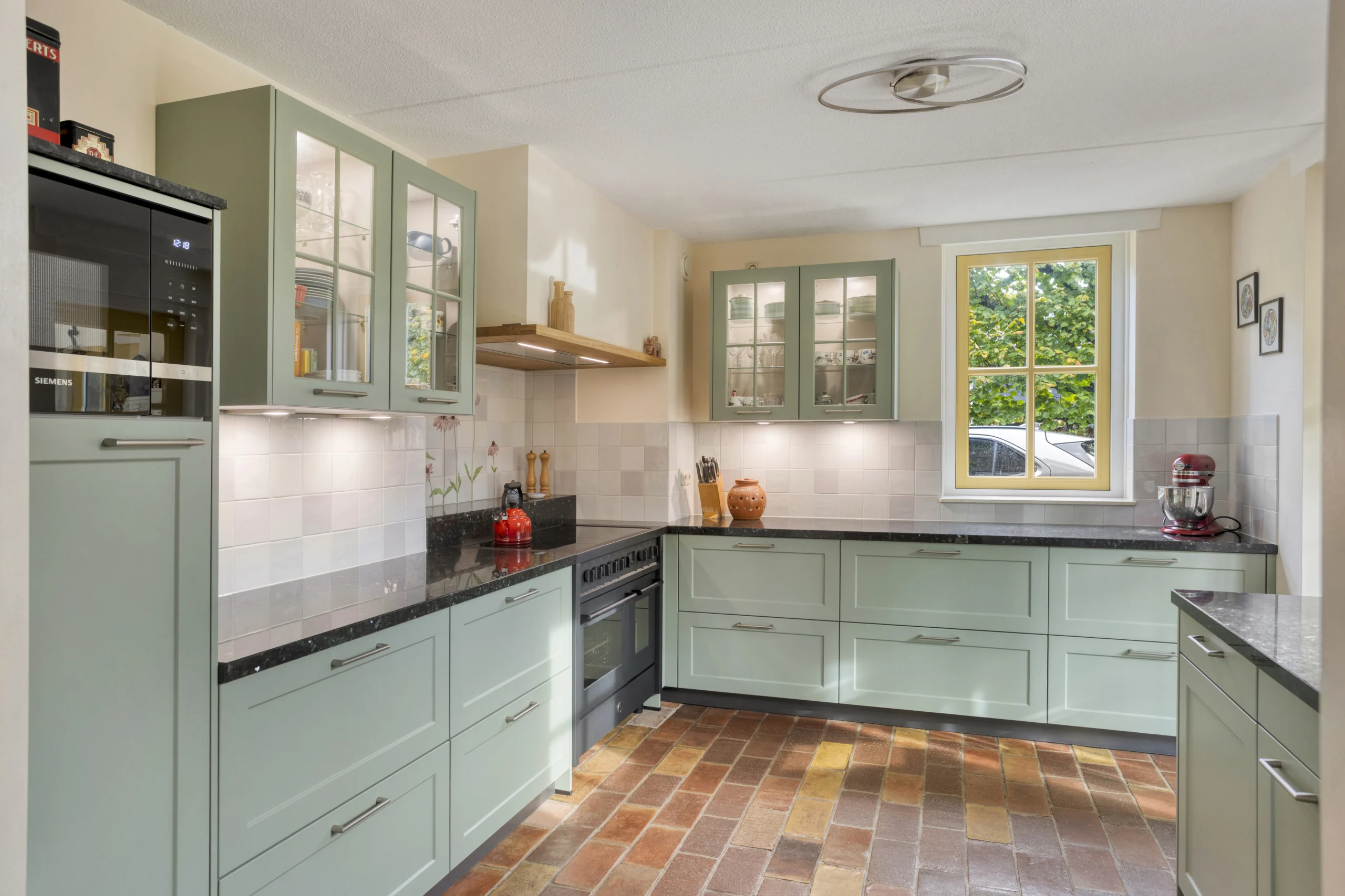 Moderne keuken met groene kasten, zwart granieten werkblad, gasfornuis, glazen deurtjes en bakstenen vloer