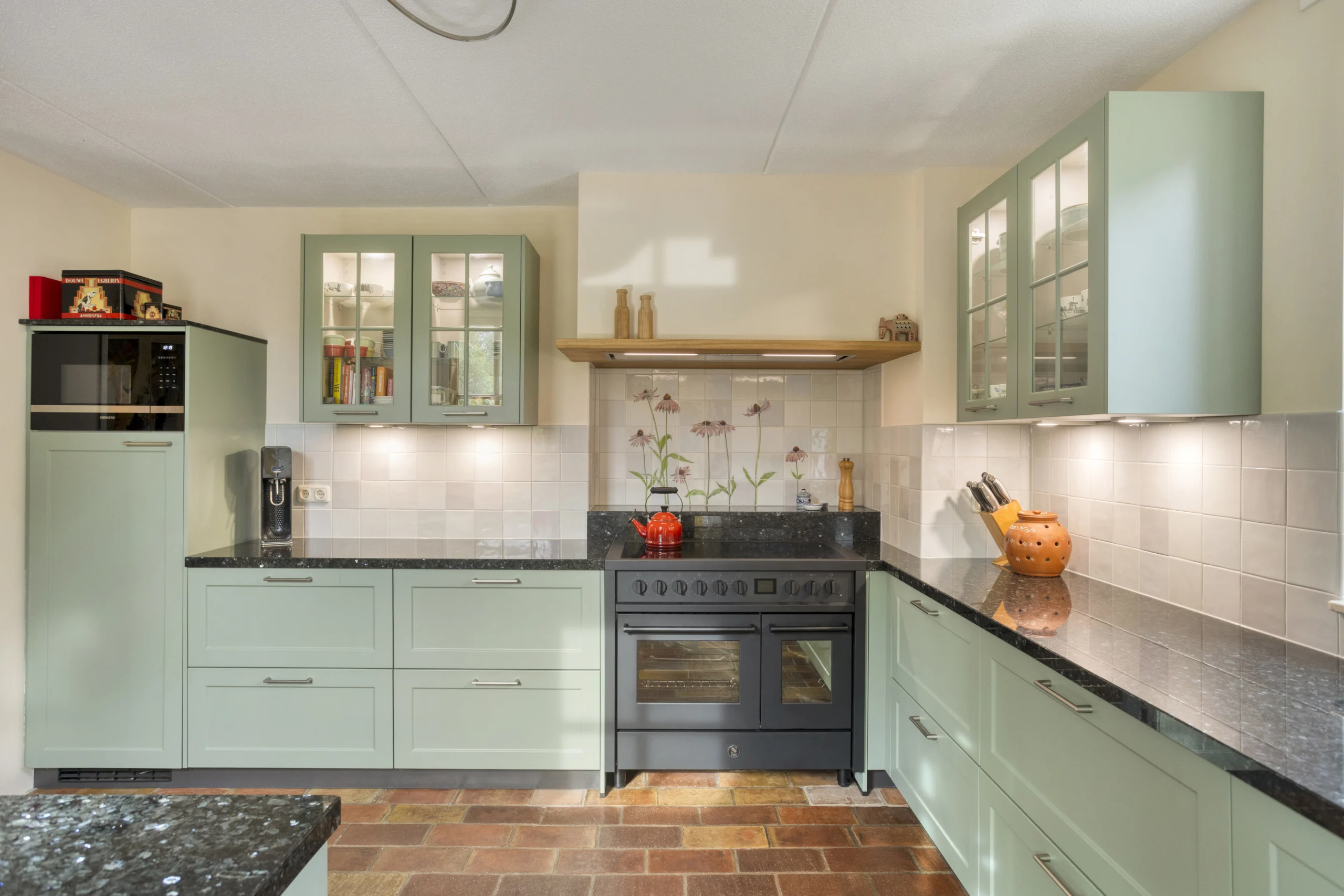 Moderne keuken met groene kasten, glazen deuren, zwart granieten werkblad en oranje kookpot.