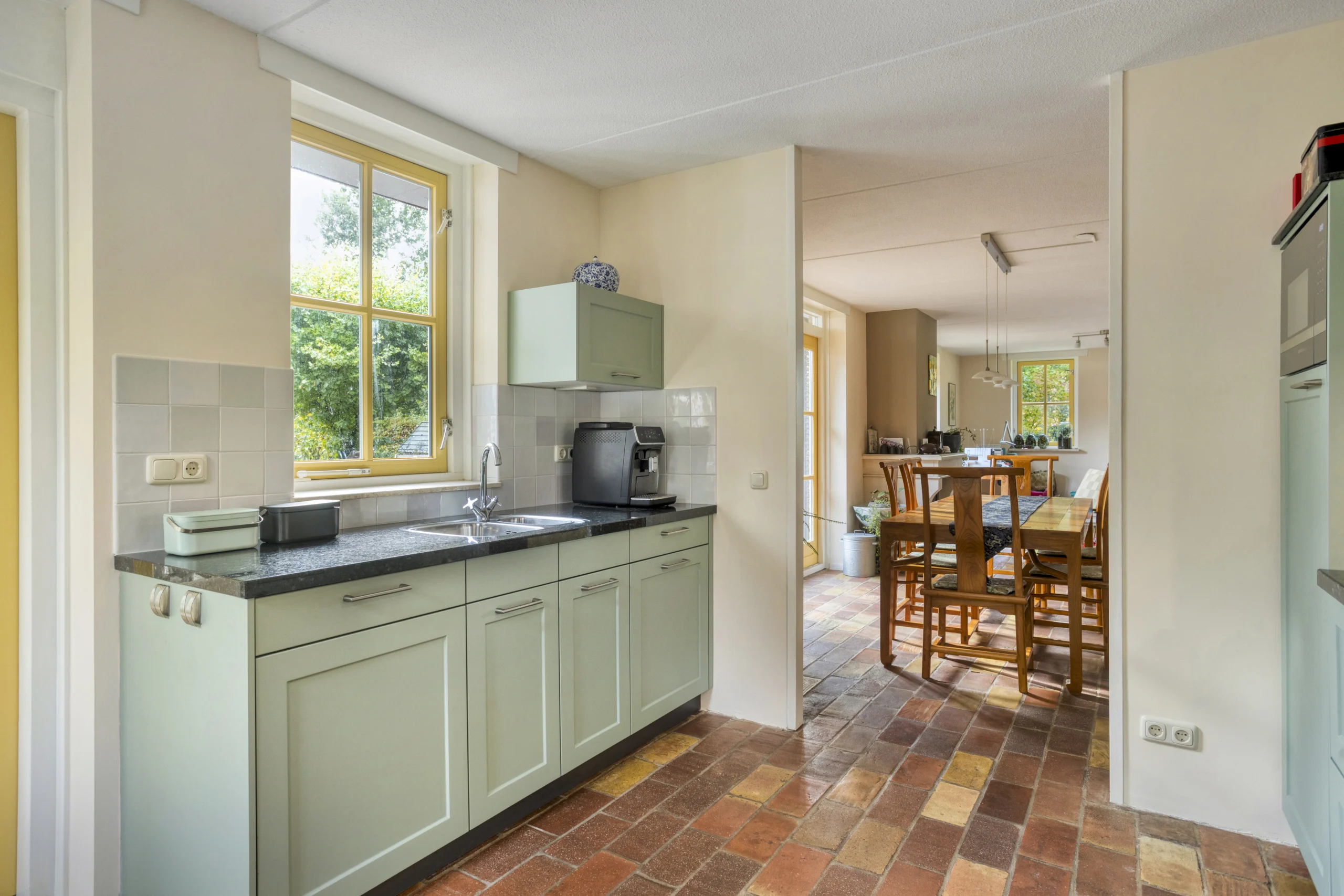 Lichte moderne keuken met lichtgroene kasten, zwart aanrecht en gele kozijnen aan groot raam