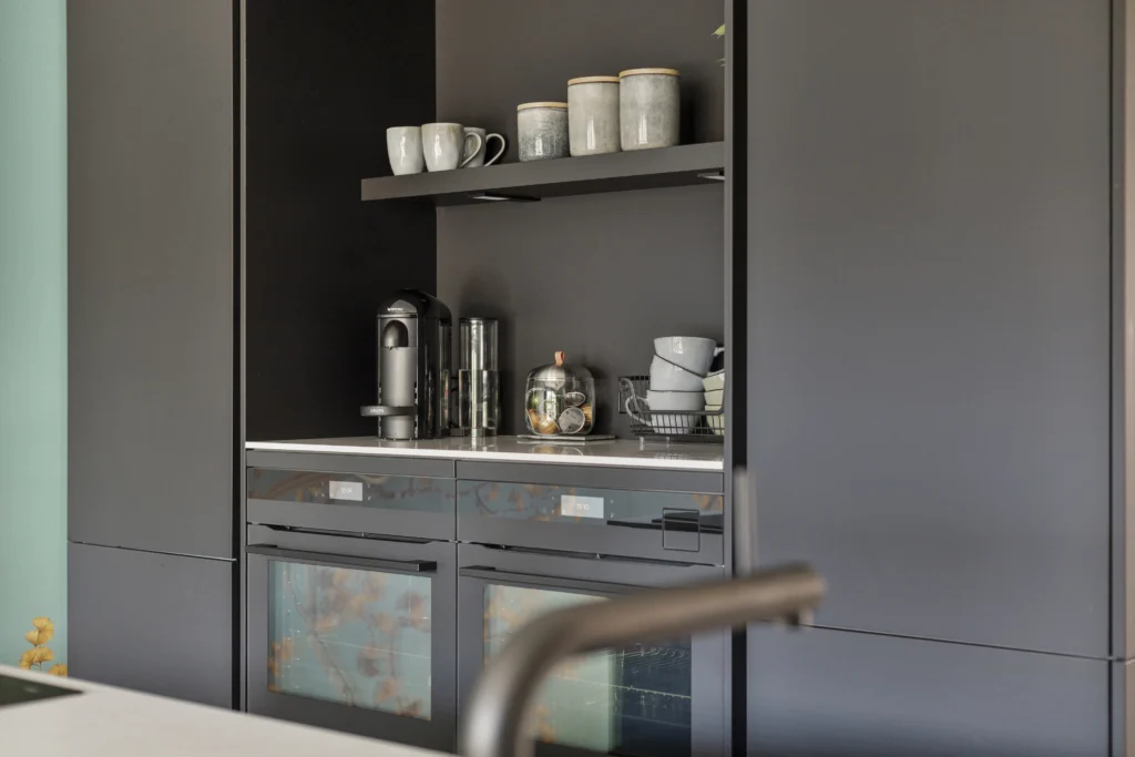 Strakke, moderne keuken met donkere kasten, grijze en witte mokken, glazen en koffiezetapparaat op plank