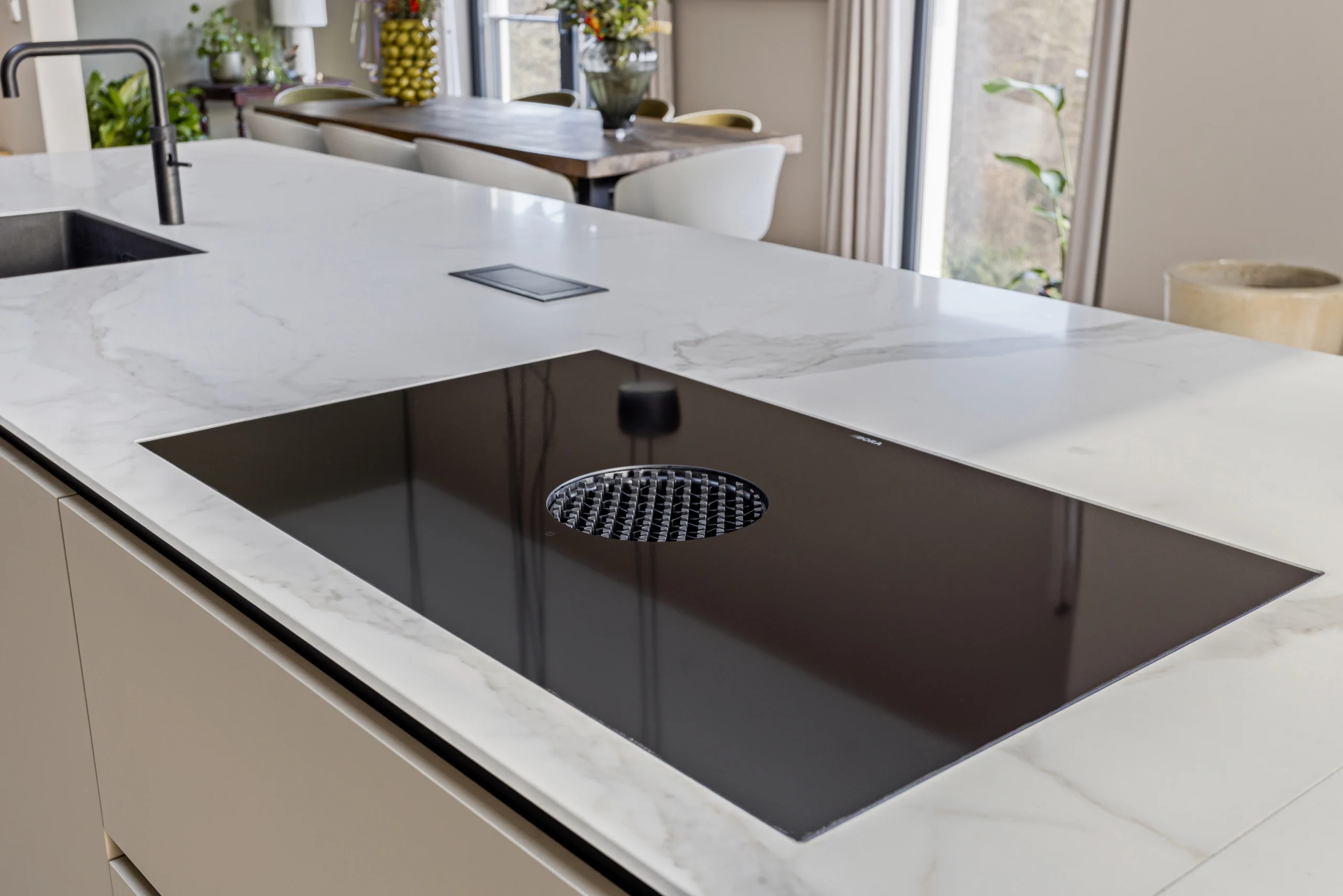 Moderne keuken met marmeren kookeiland, zwart kookoppervlak en ronde inbouwafzuiger, beige kasten en houten eettafel.