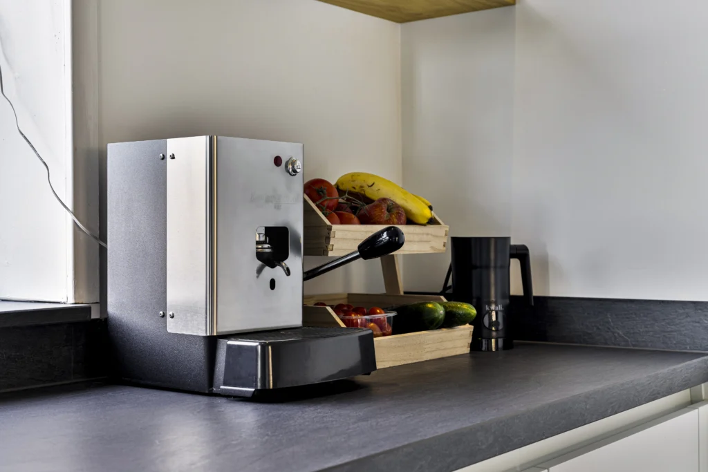 Keukenaanrecht met een moderne espressomachine, houten fruitrek en zwarte kan op een donker mat aanrecht.