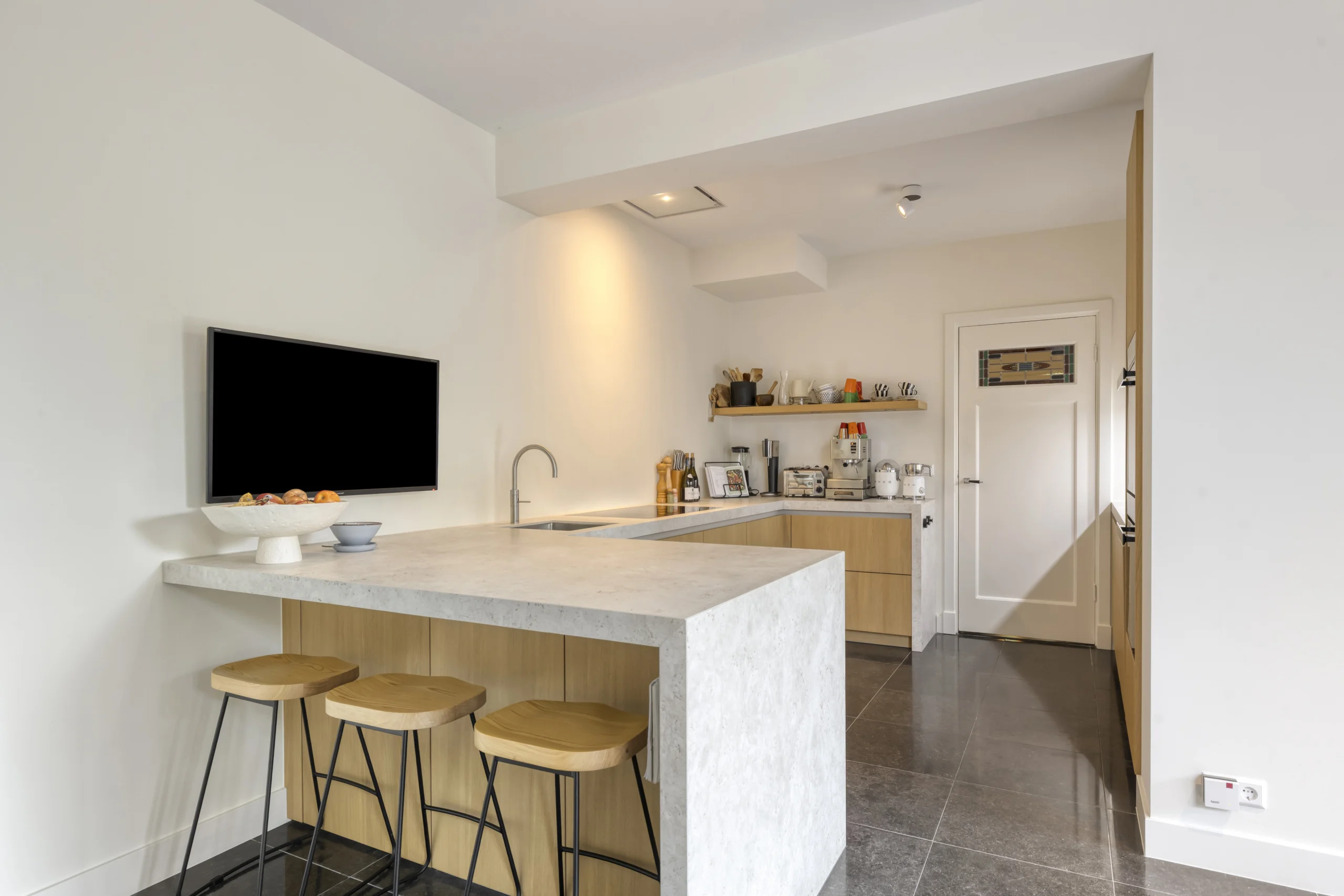 Moderne keuken met betonlook keukeneiland, houten barkrukken, licht houten kasten en donker tegelvloer