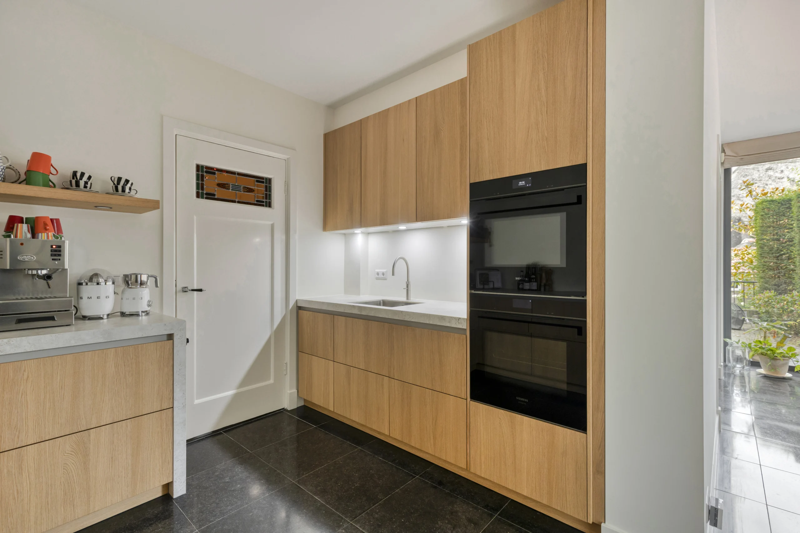 Moderne keuken met lichte houten kasten, zwart geïntegreerde oven en koffiezetapparaten naast het aanrecht.