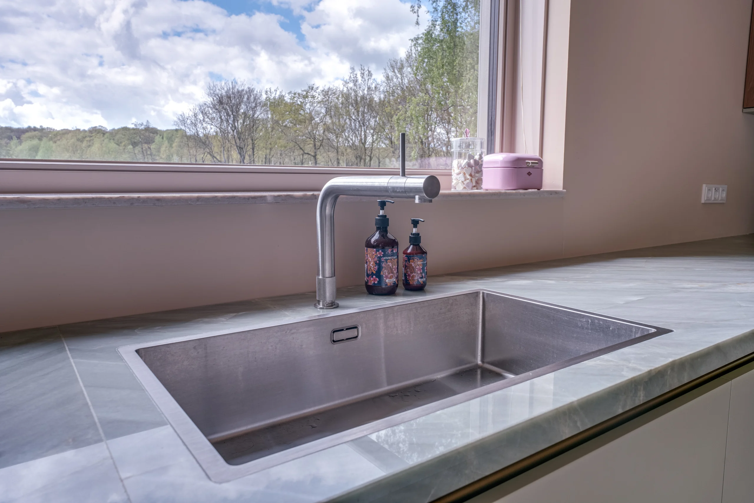 Moderne keuken met roestvrijstalen aanrecht, spoelbak, gebogen kraan en kleurrijke flessen bij groot raam.