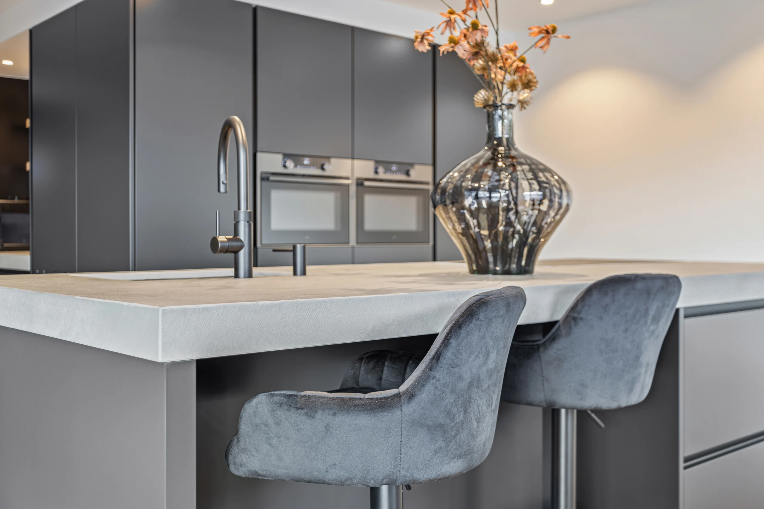 Moderne keuken met licht betonnen keukeneiland, grijze barkrukken en donkere kasten met ovens