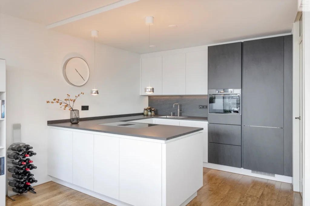 Moderne keuken met L-vormige indeling, witte en donkergrijze kastjes, hanglampen en wijnrek.